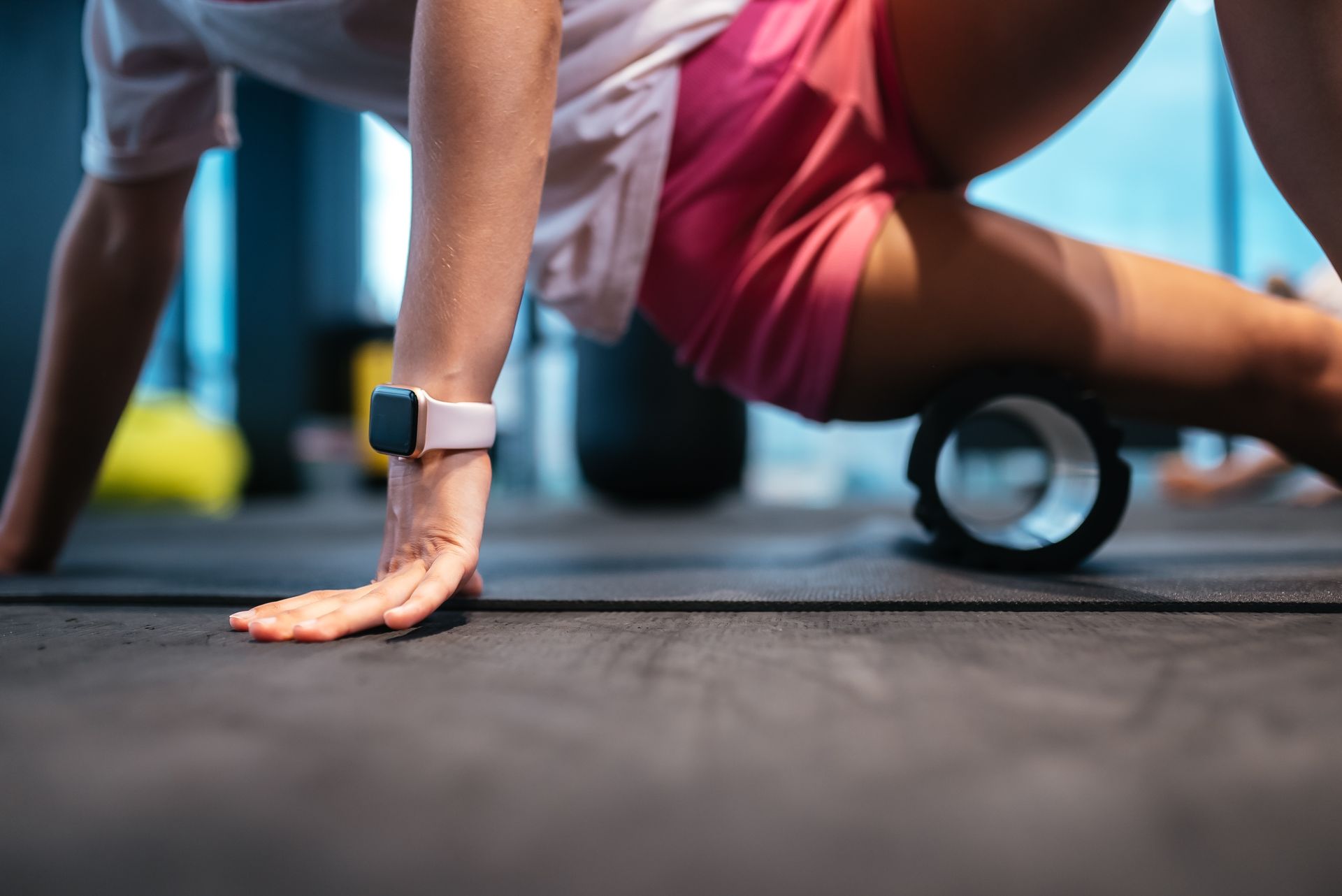 Person using a foam roller on their thigh at a gym. They are wearing pink shorts, a white shirt, and a smartwatch.