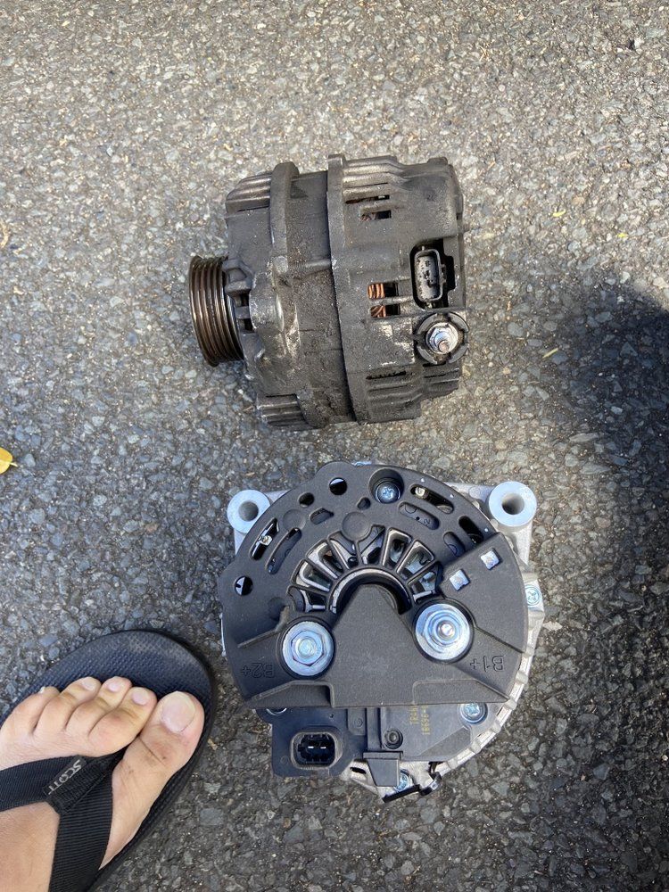 Two alternators on asphalt next to a person's foot in a flip-flop. The top one is older and worn. The bottom one is new.
