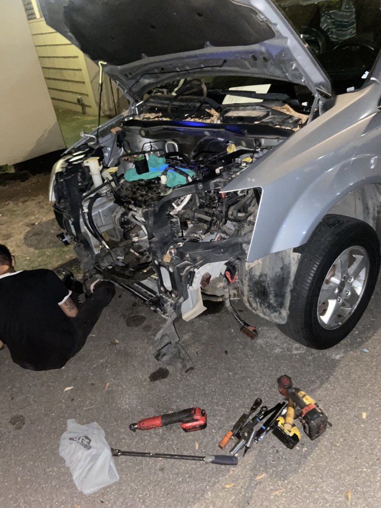 A person working on the front of a silver car with its hood open, tools scattered on the ground.