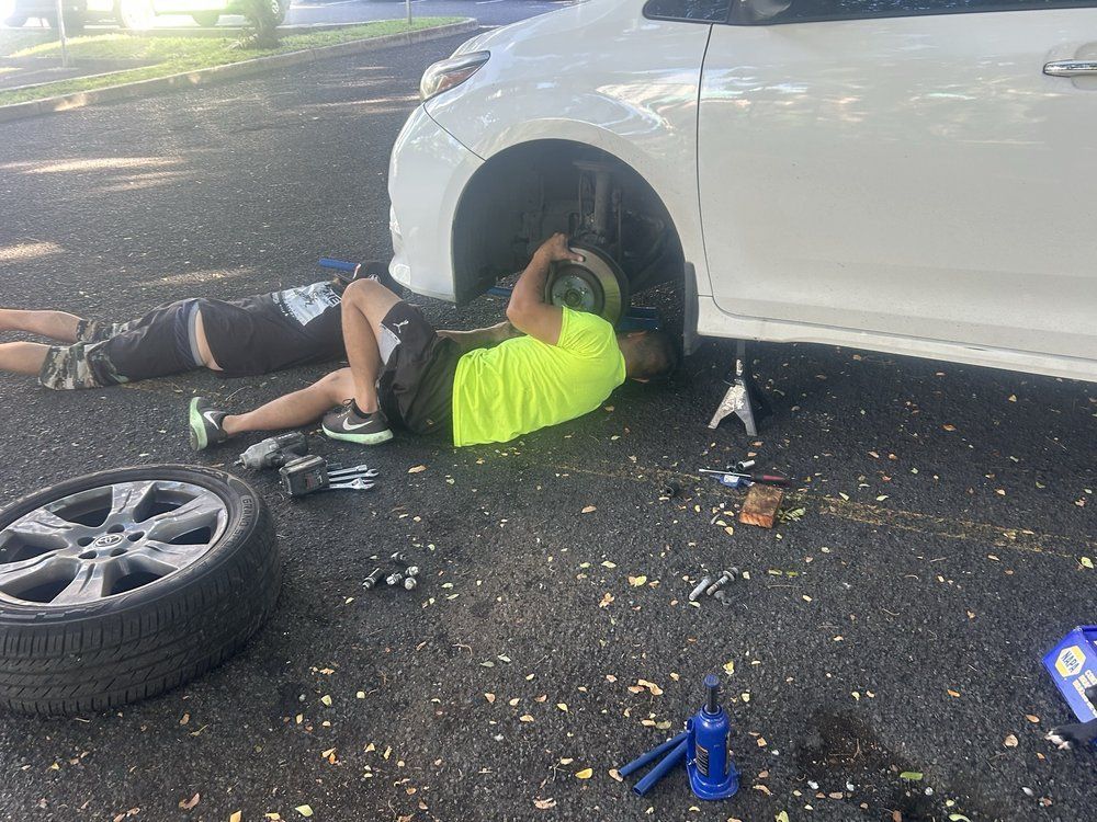 Two people work on a white car's brakes. One is under the car, the other beside it. Tools and a tire are nearby.