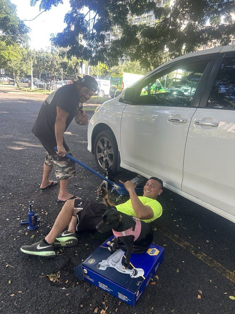 Two men changing a tire on a white minivan in a parking lot. One man is using a jack, another assisting. A dog is nearby.