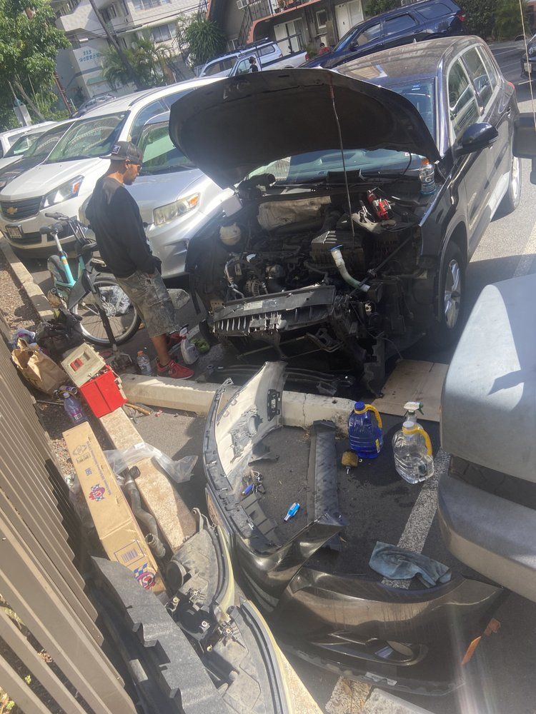 Man working on a black car with the hood up, front damaged. Tools, parts, and cardboard boxes are nearby.