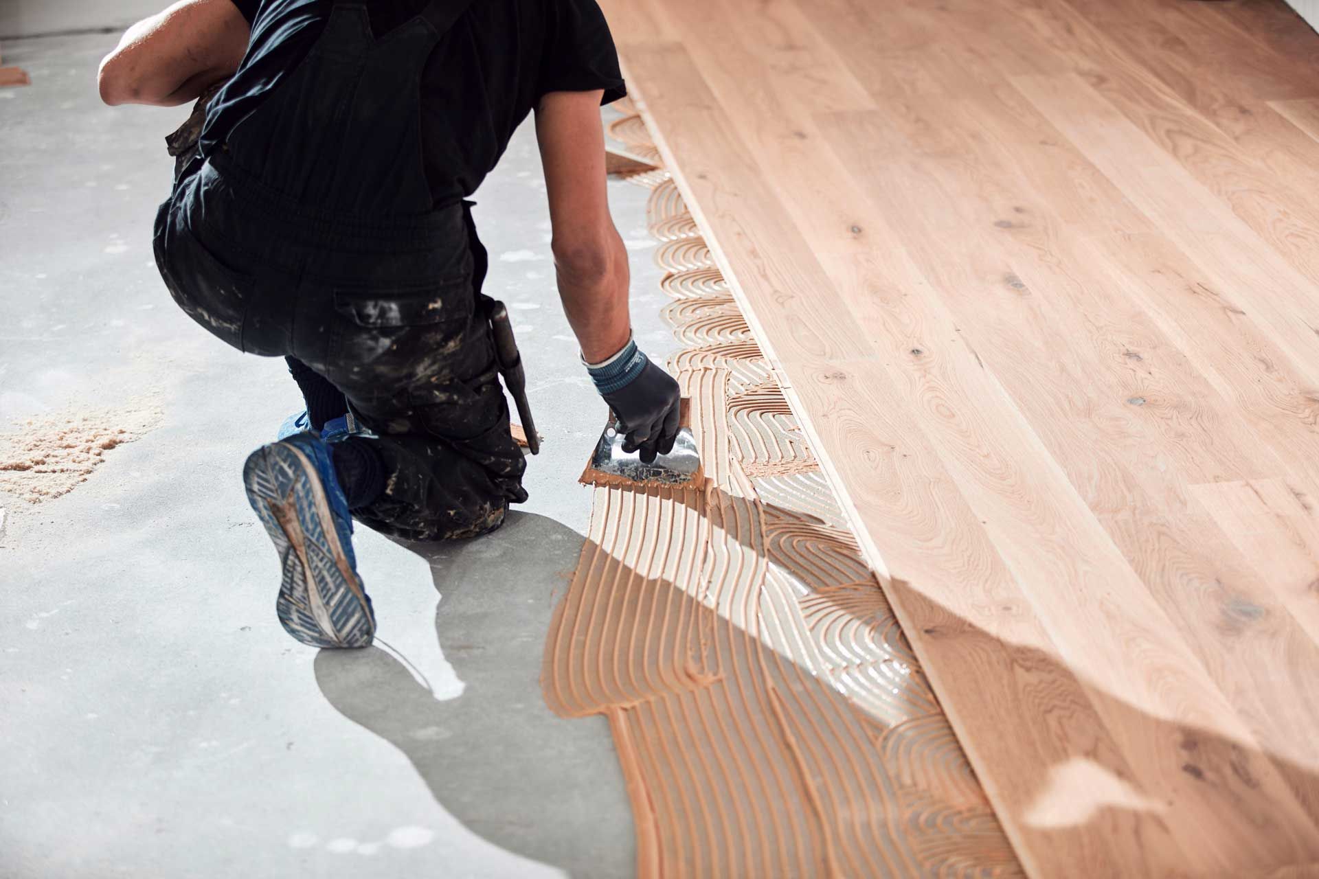 A man is kneeling down and applying glue to a wooden floor.