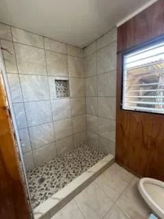 Small tiled shower stall with pebble floor, window, and wood-paneled wall beside a toilet.