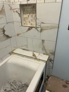 Bathroom with a bathtub and tiled wall, showing a broken wall opening and scattered debris on the floor