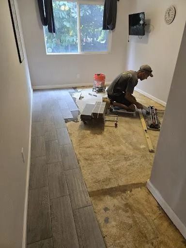 Worker installing flooring in a narrow room with a window, tools, and stacked materials on the floor