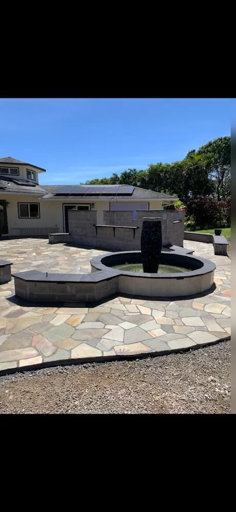 Stone patio with a circular black fountain and curved bench in a sunny backyard
