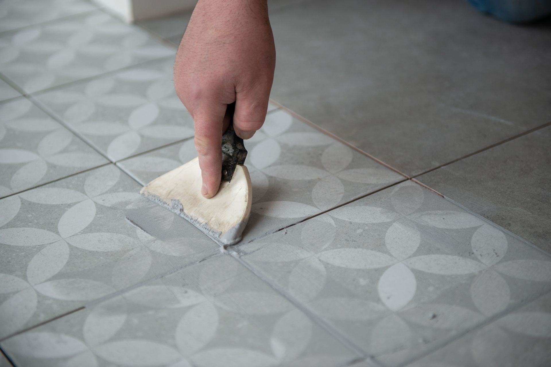 Hand spreading grout over patterned gray floor tiles with a trowel