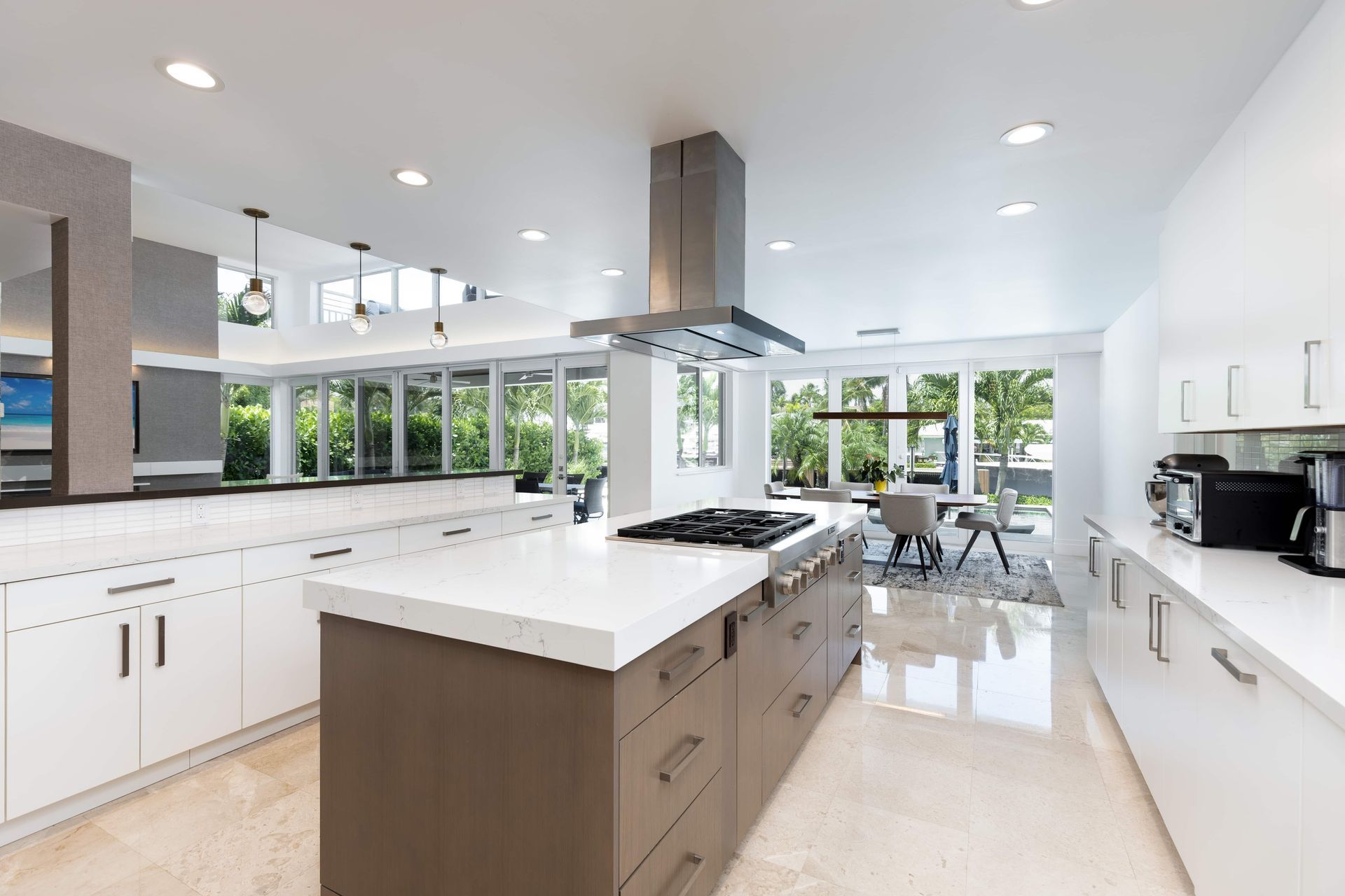 Modern white kitchen with large island, stainless range hood, and bright windows overlooking greenery