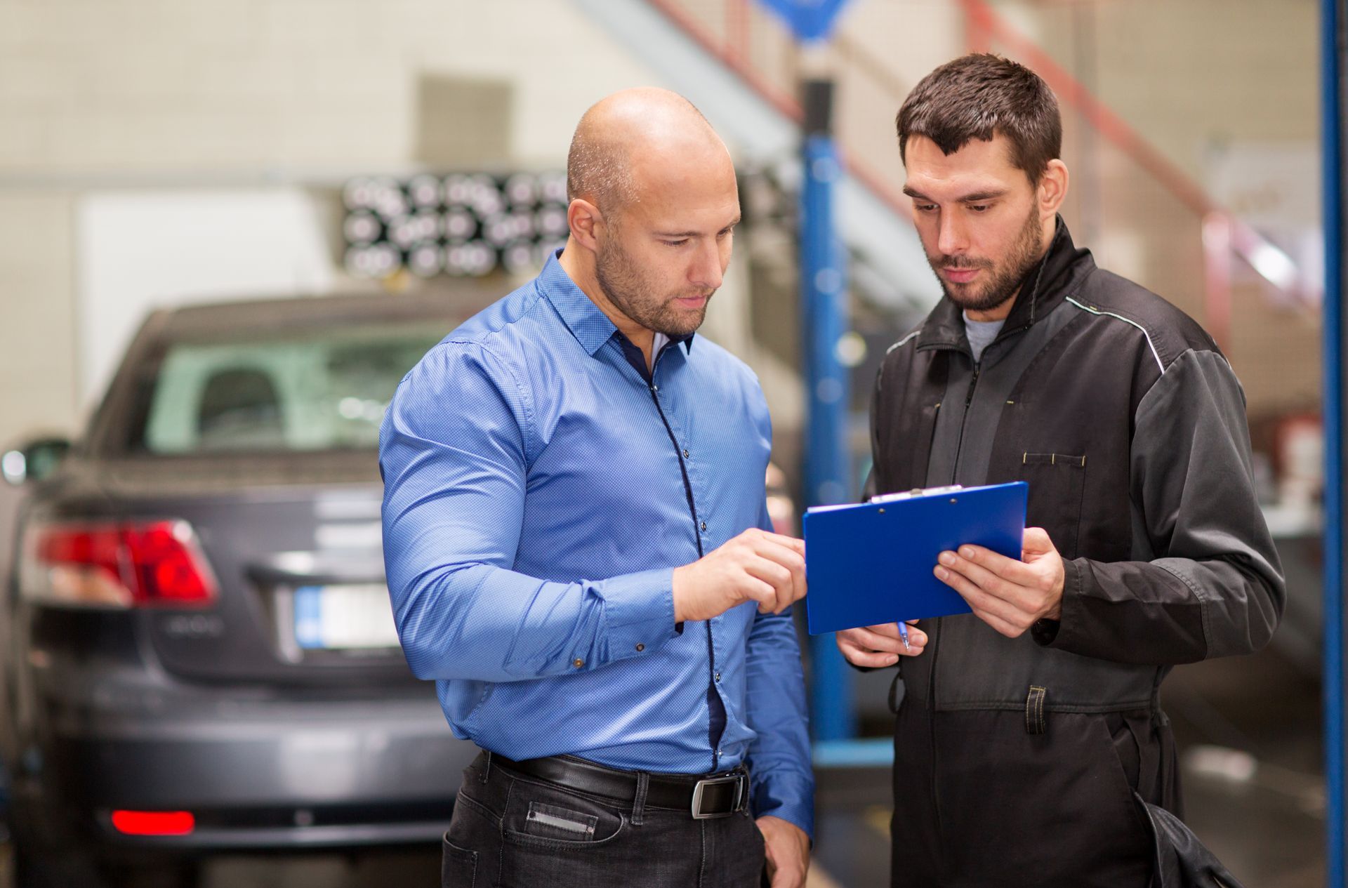 Man in blue shirt reviews paperwork with mechanic in a garage; car in the background.