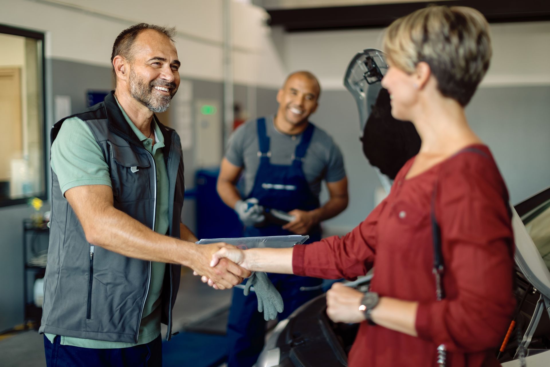 Mechanic shaking hands with a woman in a car repair shop; another mechanic in the background.
