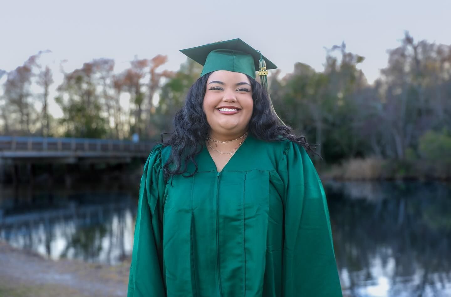 Person in green graduation gown and cap smiling by water, trees, and a bridge.