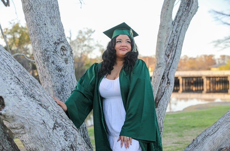 Woman in green graduation gown and cap poses in tree with a lake in the background.