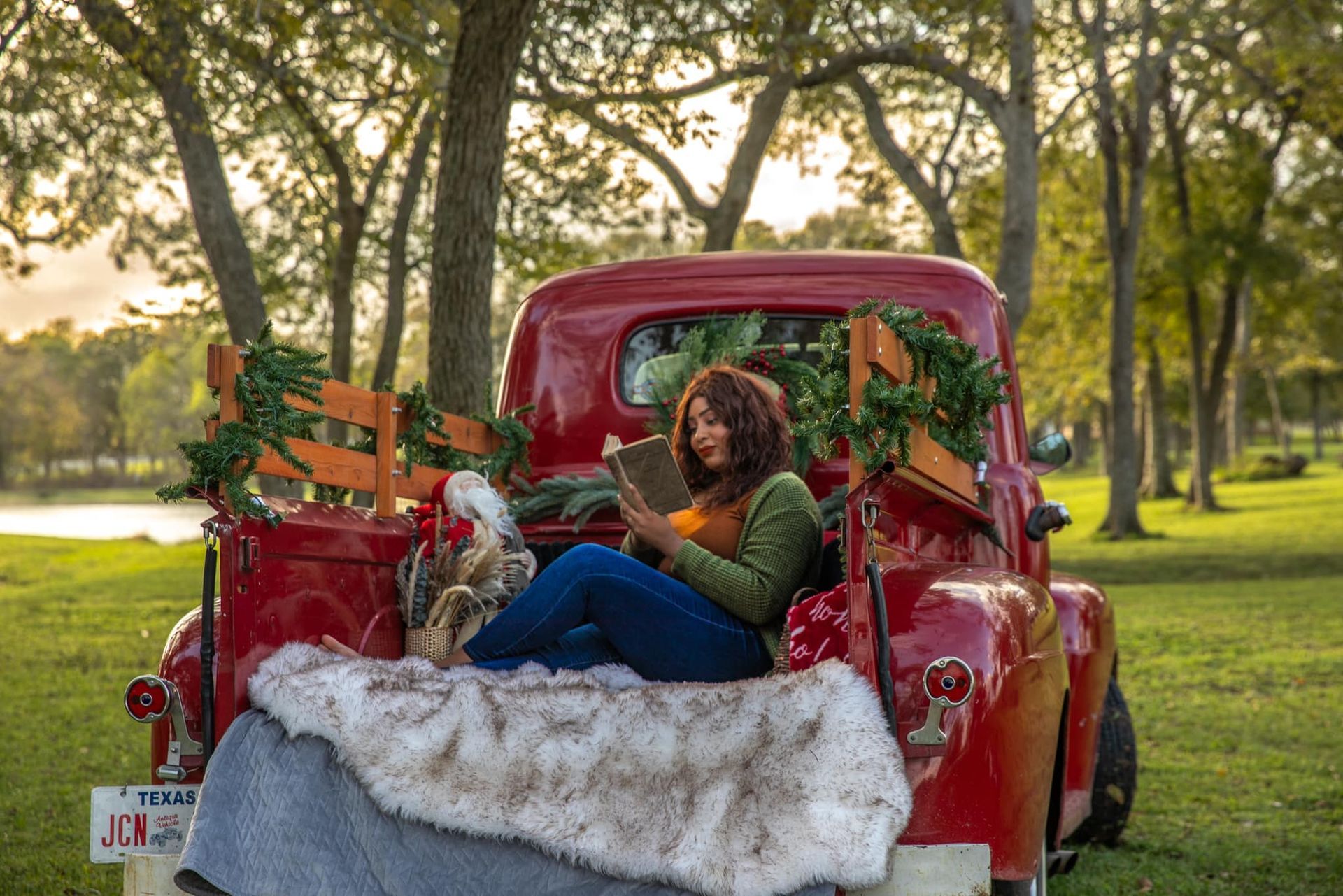 Woman reading in the back of a red truck decorated with greenery, seated on a fur blanket. Outdoors, trees in background.