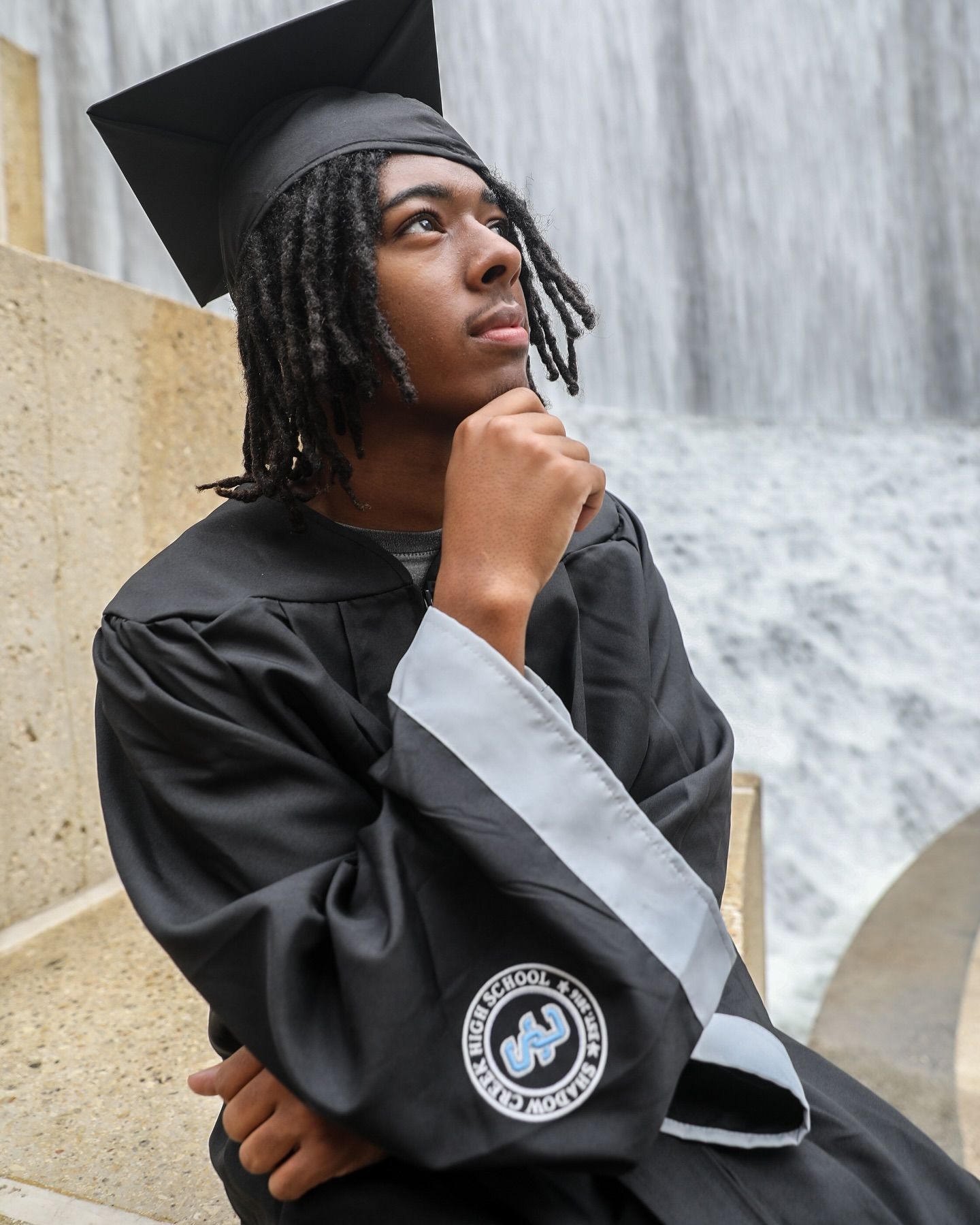 Person in graduation attire looks up pensively, waterfall backdrop.
