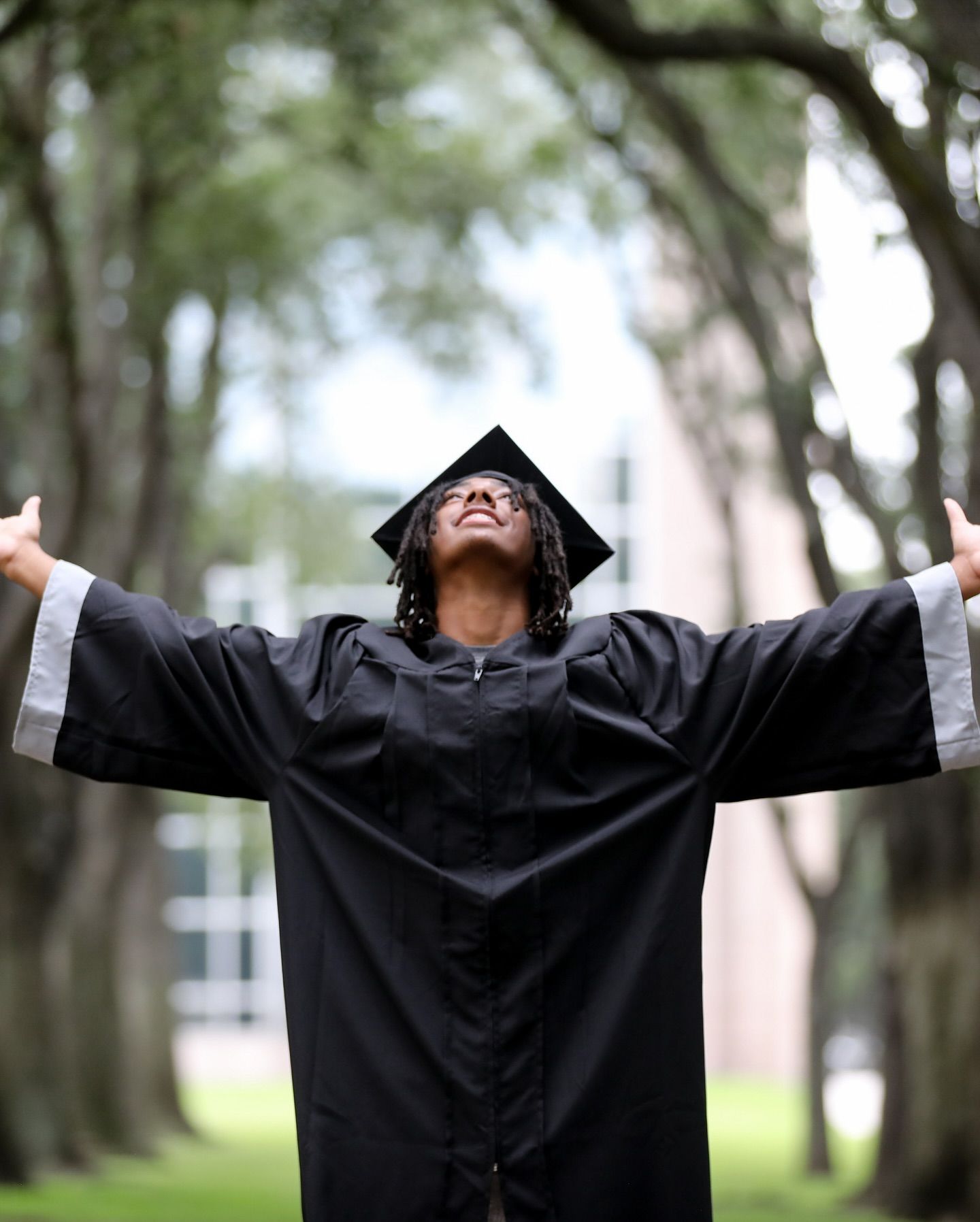 Person in graduation gown with arms raised, looking up, outdoors.