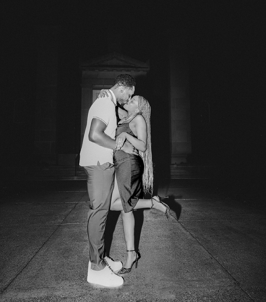Couple kissing in front of a dark building entrance. Woman lifts one leg. Black and white.