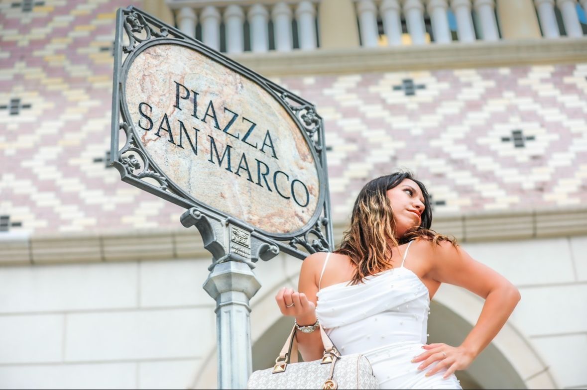 Woman in white dress posing by a Piazza San Marco sign, with patterned building in background.