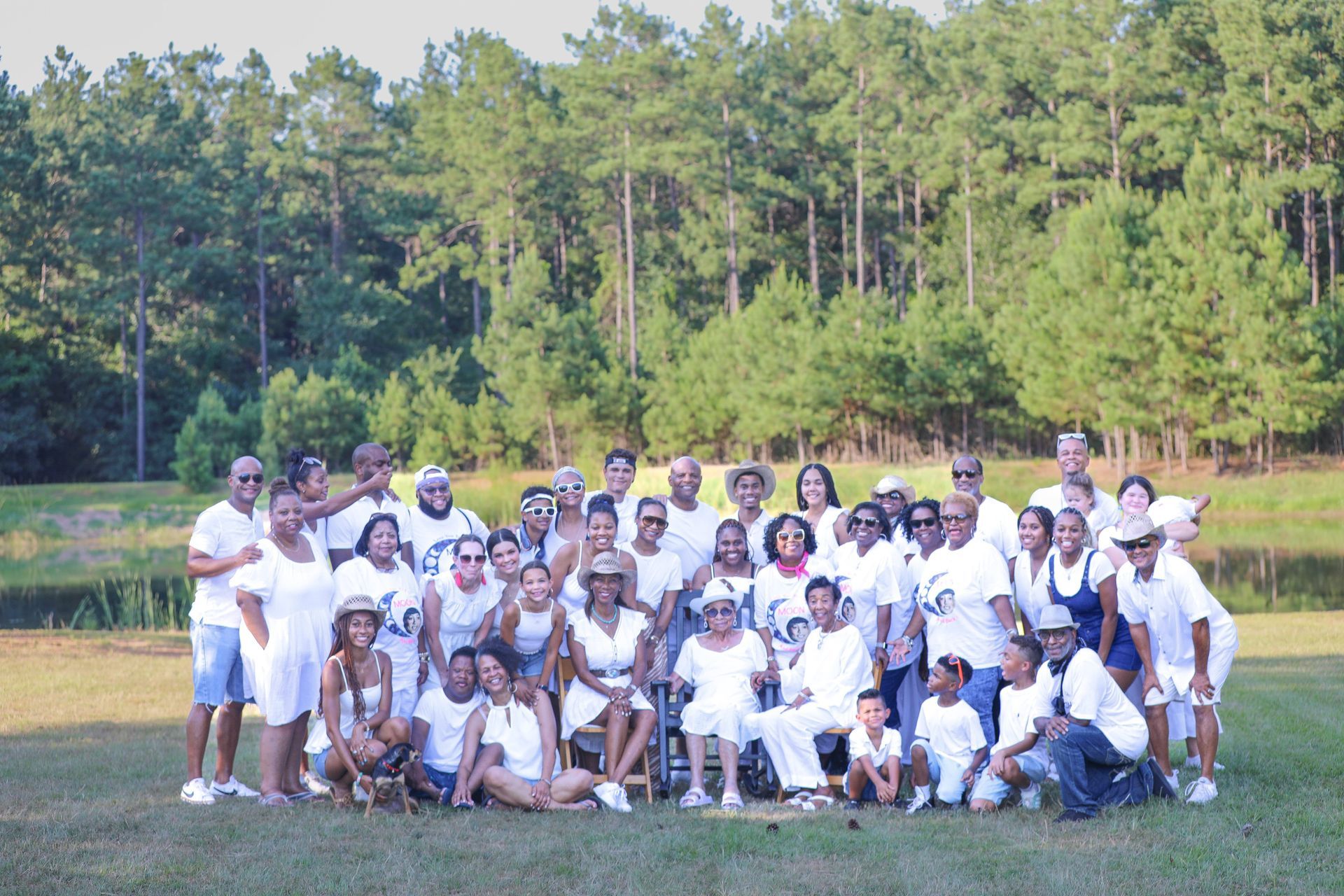 Large group of people in white clothing pose on a grassy field in front of a forest and pond.