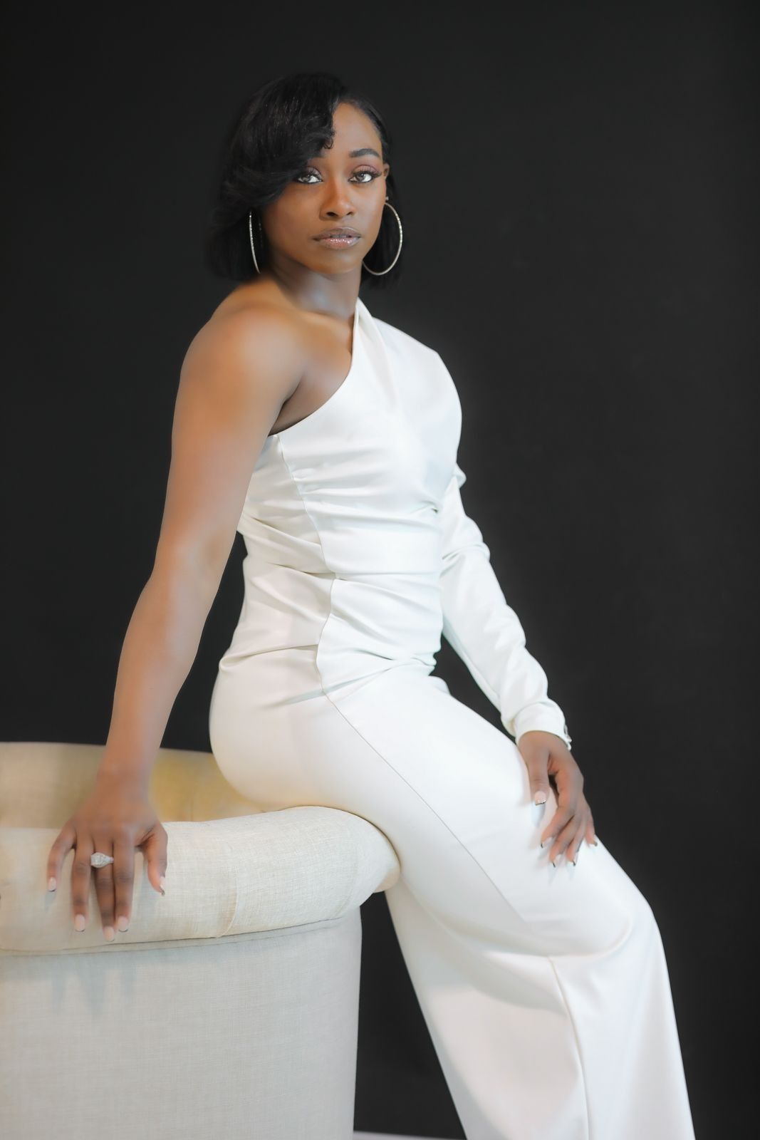 Woman in white one-shoulder jumpsuit sits on cream-colored structure, gold hoop earrings, black background.