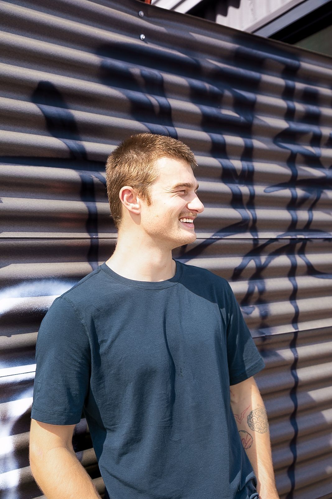 Man in dark blue shirt smiles, standing against corrugated metal wall with graffiti.