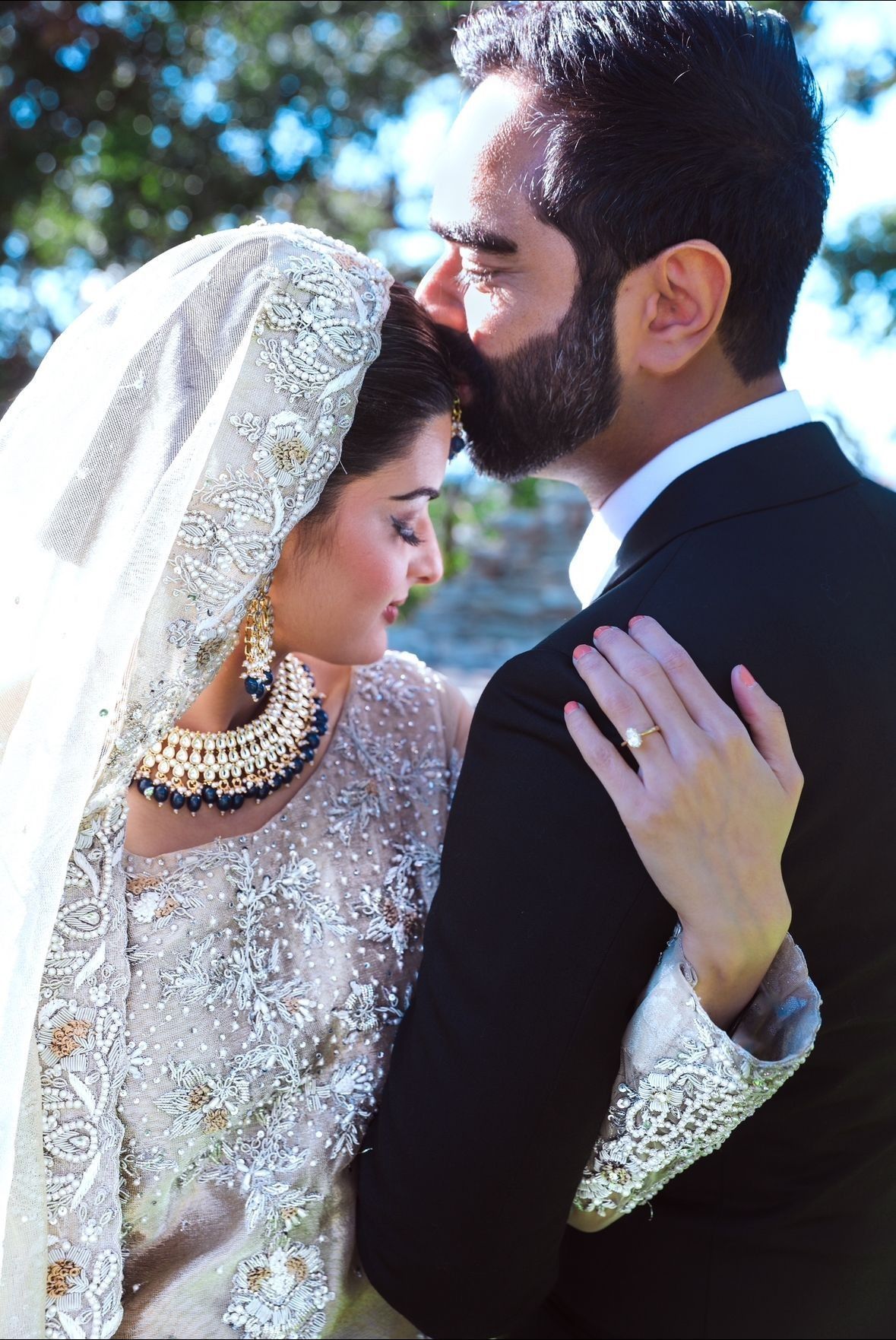 Bride in embellished veil and jewelry, groom in suit, embracing; outdoor setting.