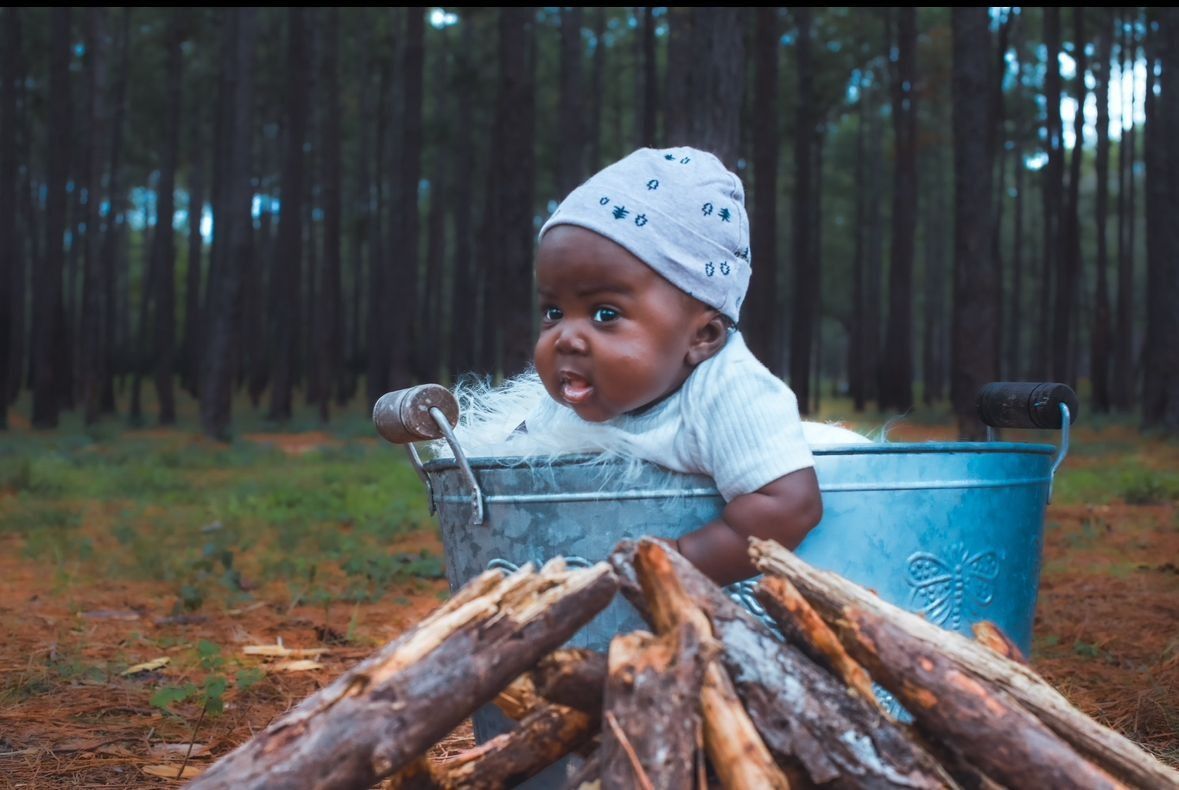A baby in a metal tub near a pile of logs in a forest. Baby looks at the camera.
