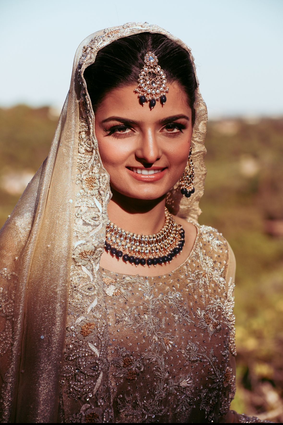 Woman in gold wedding attire, smiling; wears veil, jewelry. Outdoor setting, soft focus.