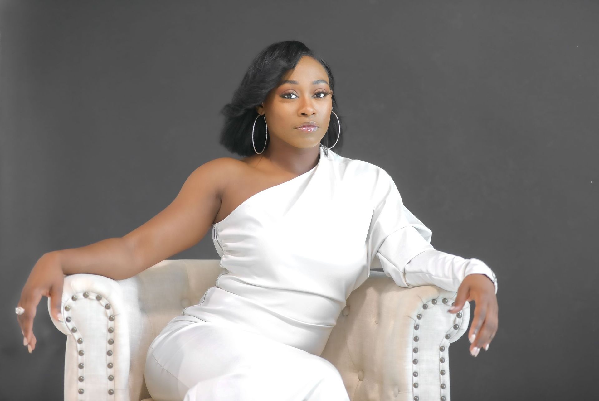 Woman in white one-shoulder dress sits in an off-white armchair against a dark gray backdrop.