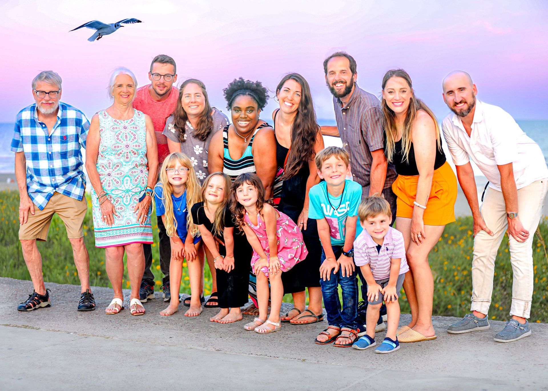 Large group poses for photo outdoors at sunset, seaside; mix of adults and children smiling.