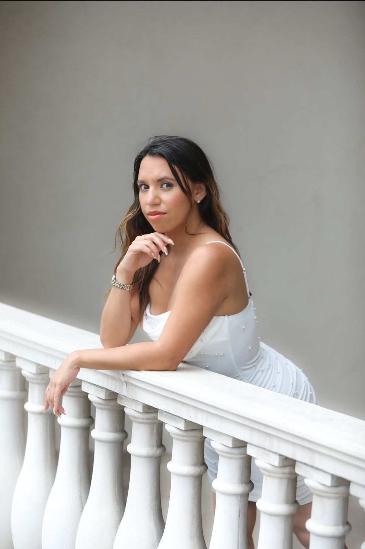 Woman in white dress leans on a white railing, gazing at the camera.
