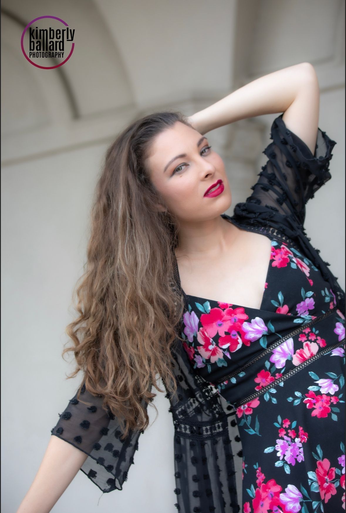 Woman in floral dress, arm raised, looking up. Curly hair, red lipstick, wearing sheer black jacket.