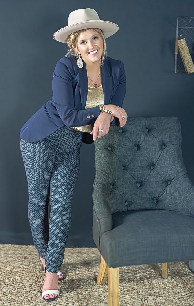 Woman in navy blazer and patterned pants, leaning on a gray chair, smiling, wearing a hat.