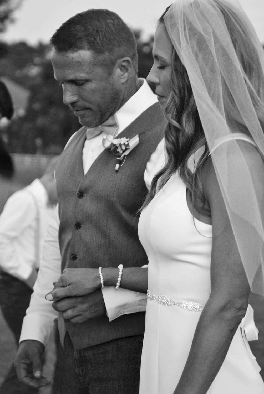 Bride and groom holding hands, walking together; man in vest, woman in wedding dress, outside.