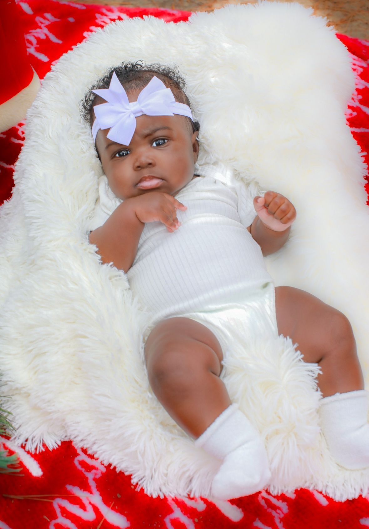 Baby in white outfit and bow, lying on a fuzzy white blanket atop a red patterned surface.