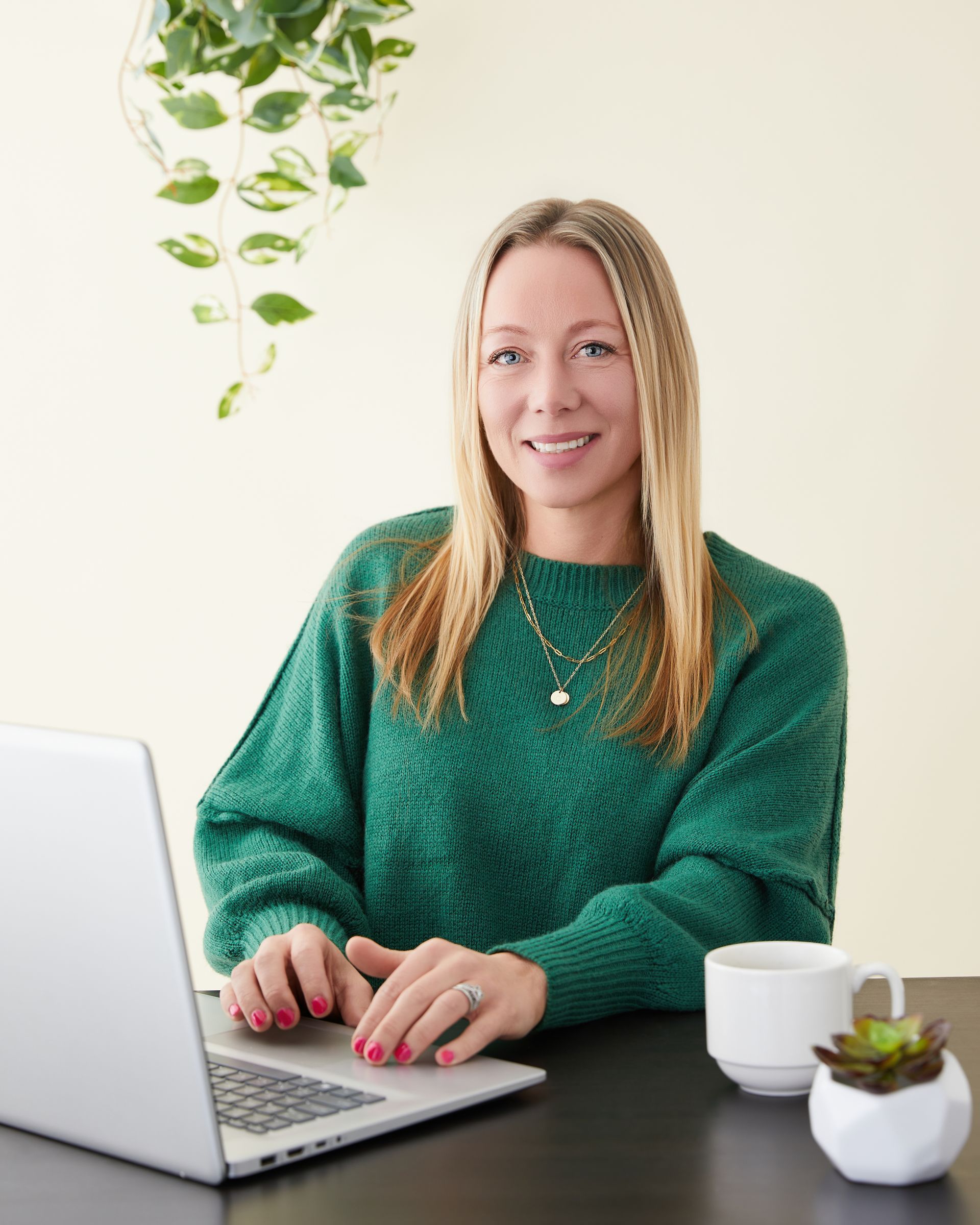 Woman with blonde hair smiles, leans against a dark metal wall, wearing a black blazer and teal shirt outdoors.