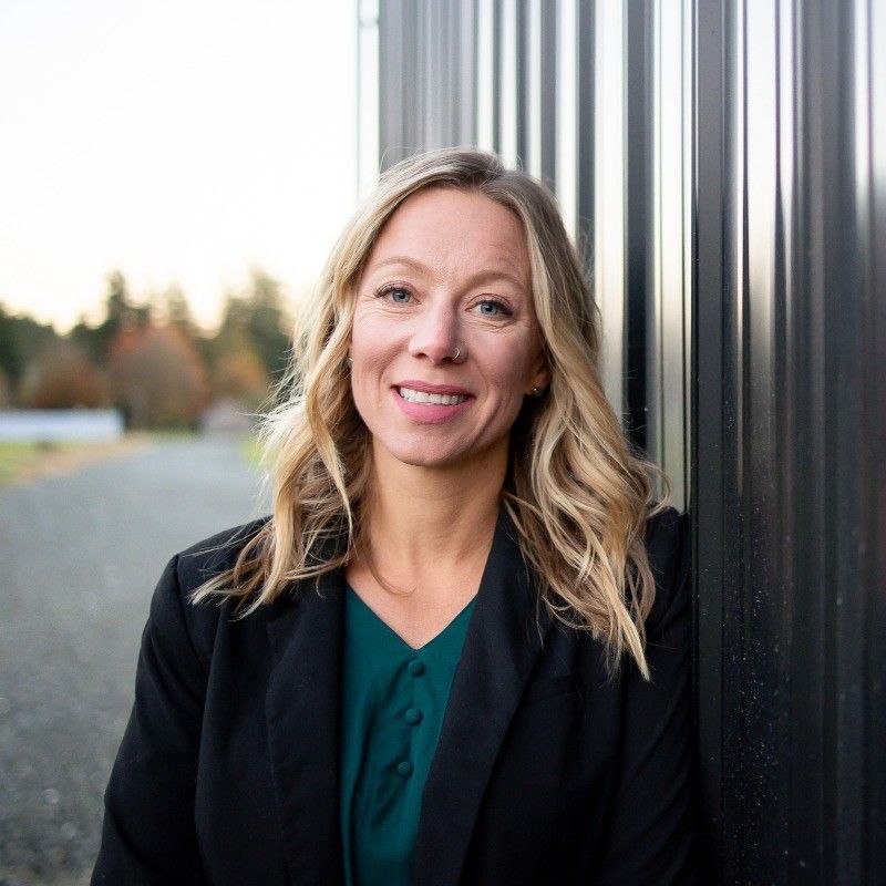 Woman with blonde hair smiles, leans against a dark metal wall, wearing a black blazer and teal shirt outdoors.