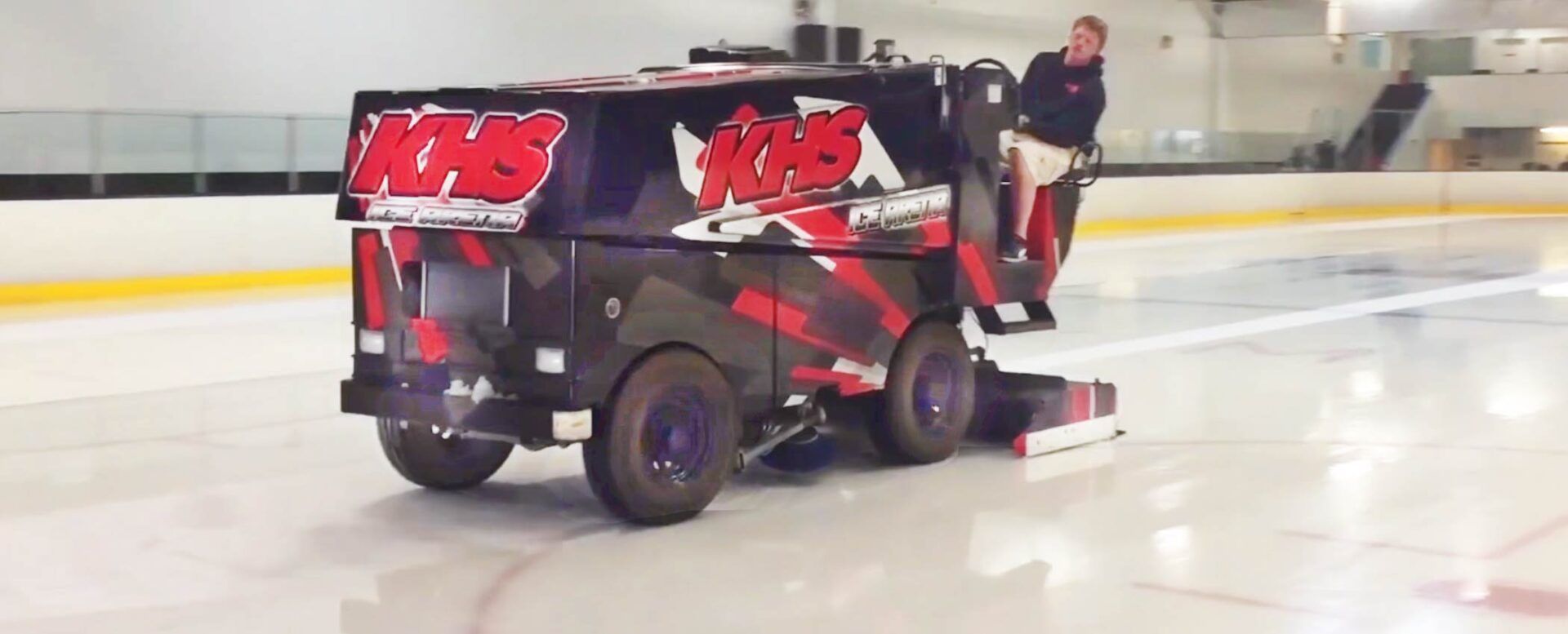 An ice resurfacer with black and red branding drives across an indoor ice rink while a person operates it from the back.