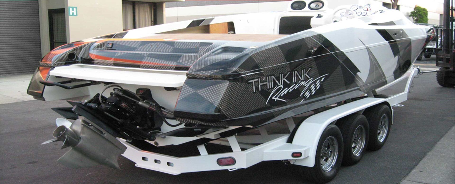 A high-performance speedboat with a black, white, and gray geometric pattern, loaded on a white triple-axle trailer.