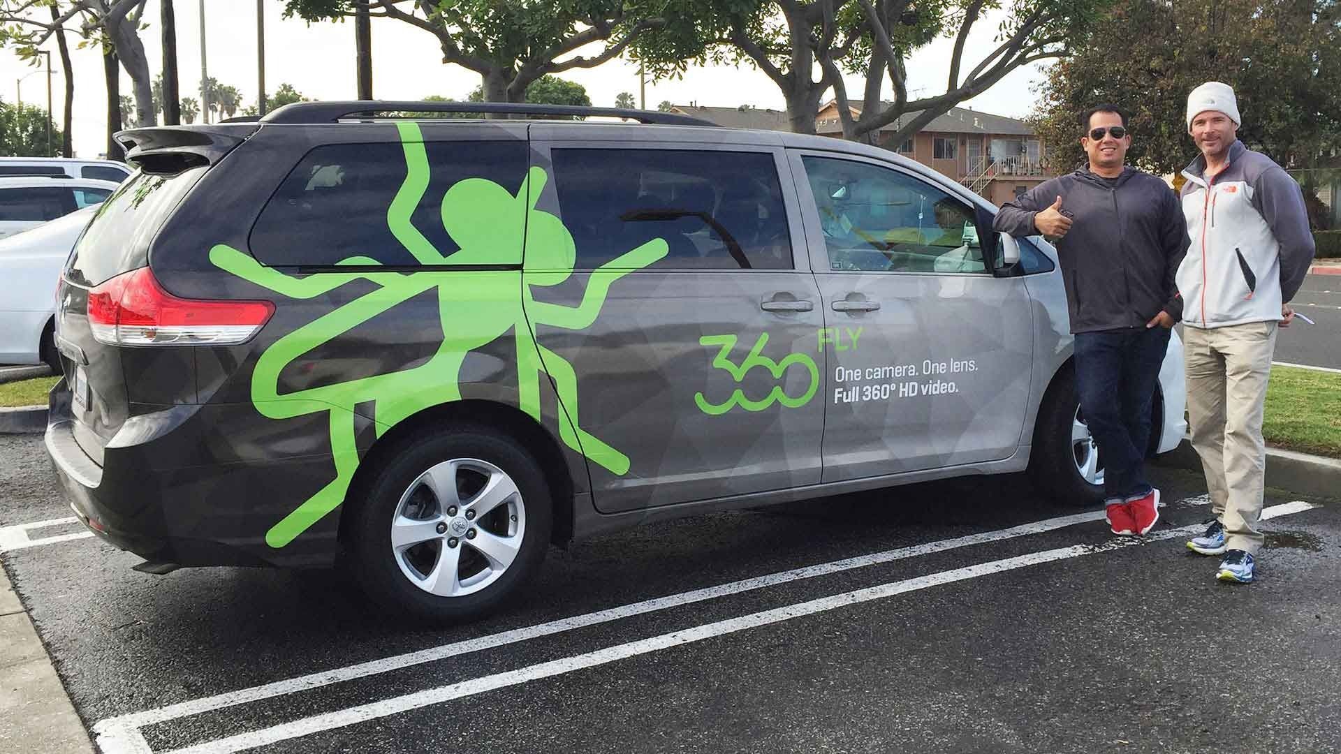 Two people stand next to a dark gray minivan with a green ant graphic and the 