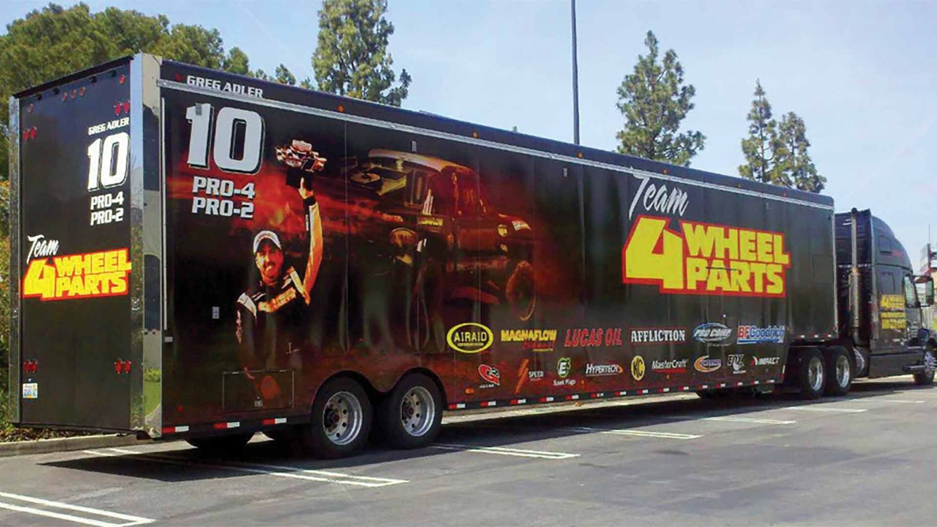 A 4 Wheel Parts semi-trailer parked outdoors, featuring a race car driver, number 10 branding, and sponsor logos.