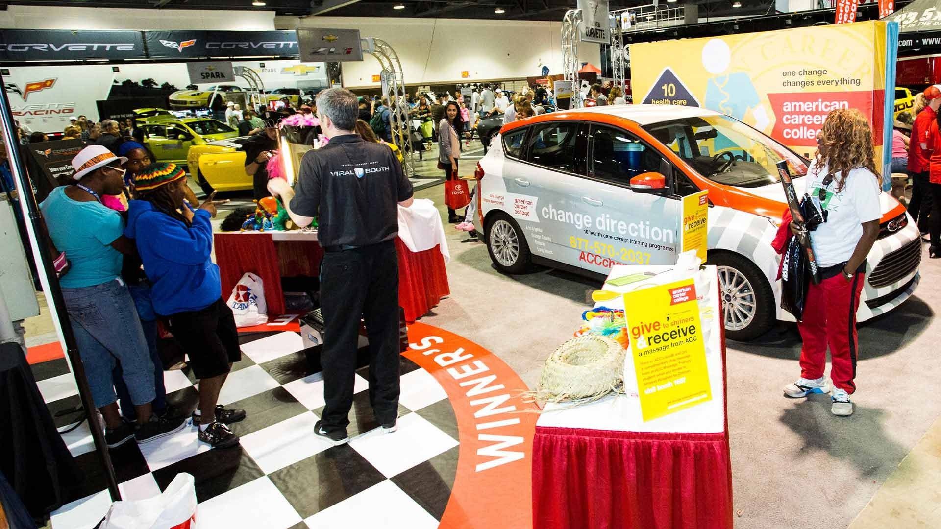 People gather at a trade show booth featuring a silver display car on a black-and-white checkered floor.