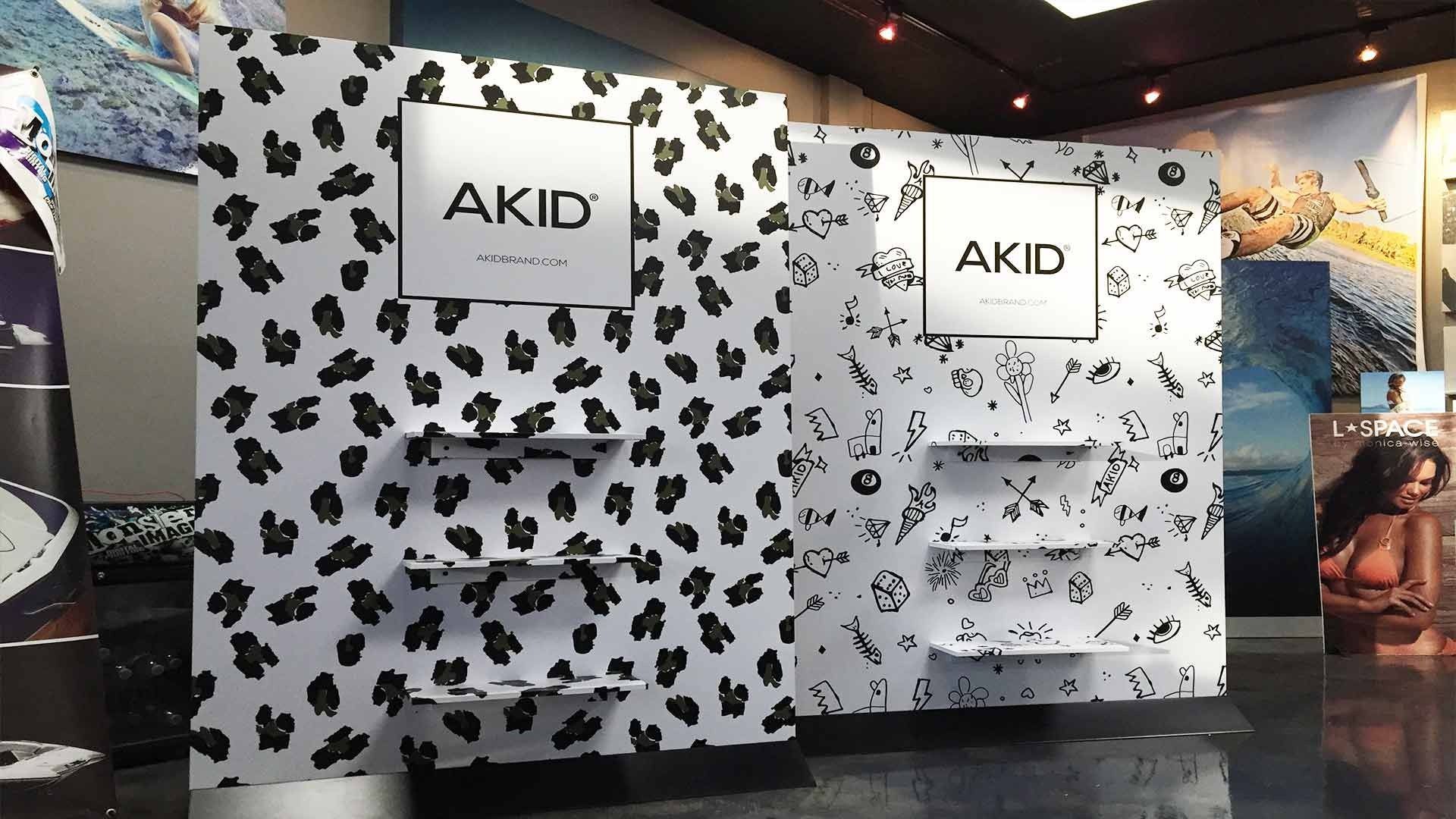 Two side-by-side retail displays with the AKID logo, featuring black and white patterned backgrounds and tiered shelves.