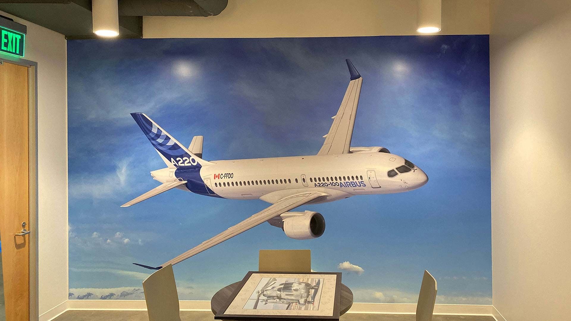A wall mural of an Airbus A220 aircraft flying against a blue sky, set in a room with a small table and chairs.
