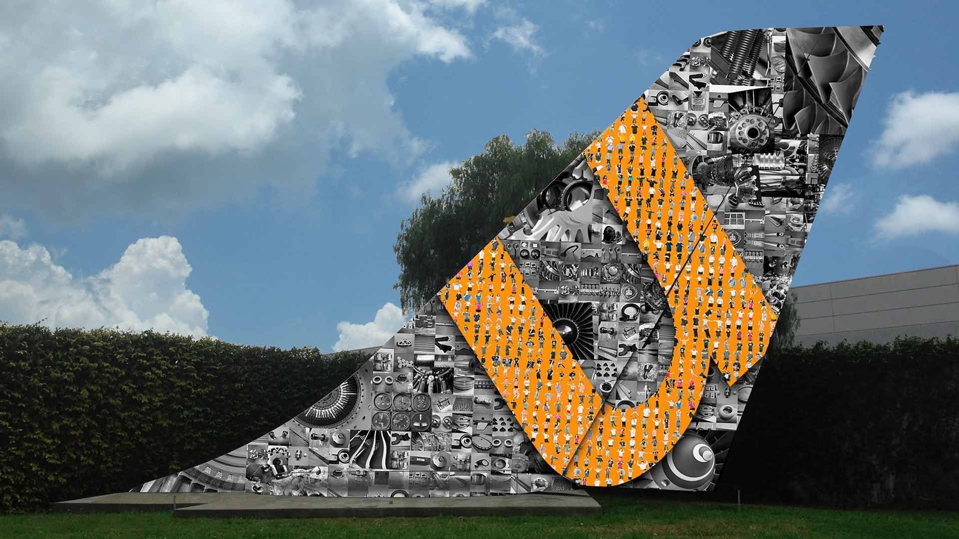 A vertical tail fin sculpture featuring a collage of black-and-white mechanical parts and a large orange 