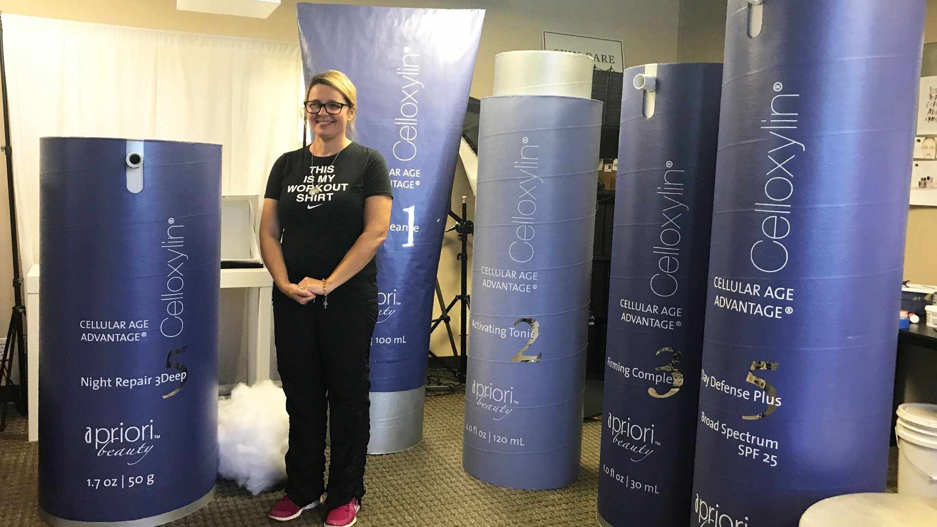 A person stands beside five oversized purple skincare product replicas in a professional office or studio setting.