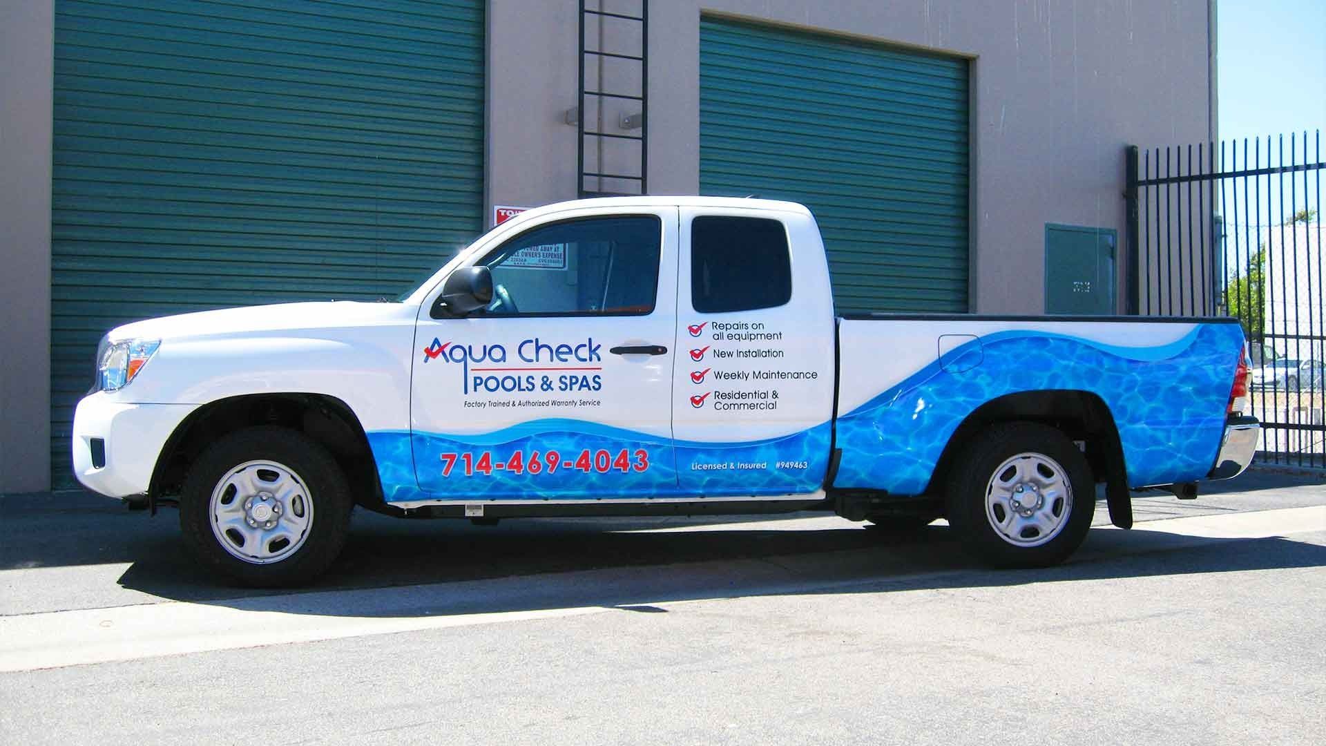 White pickup truck branded for Aqua Chek Pools & Spas, featuring blue water graphics parked in front of a warehouse.