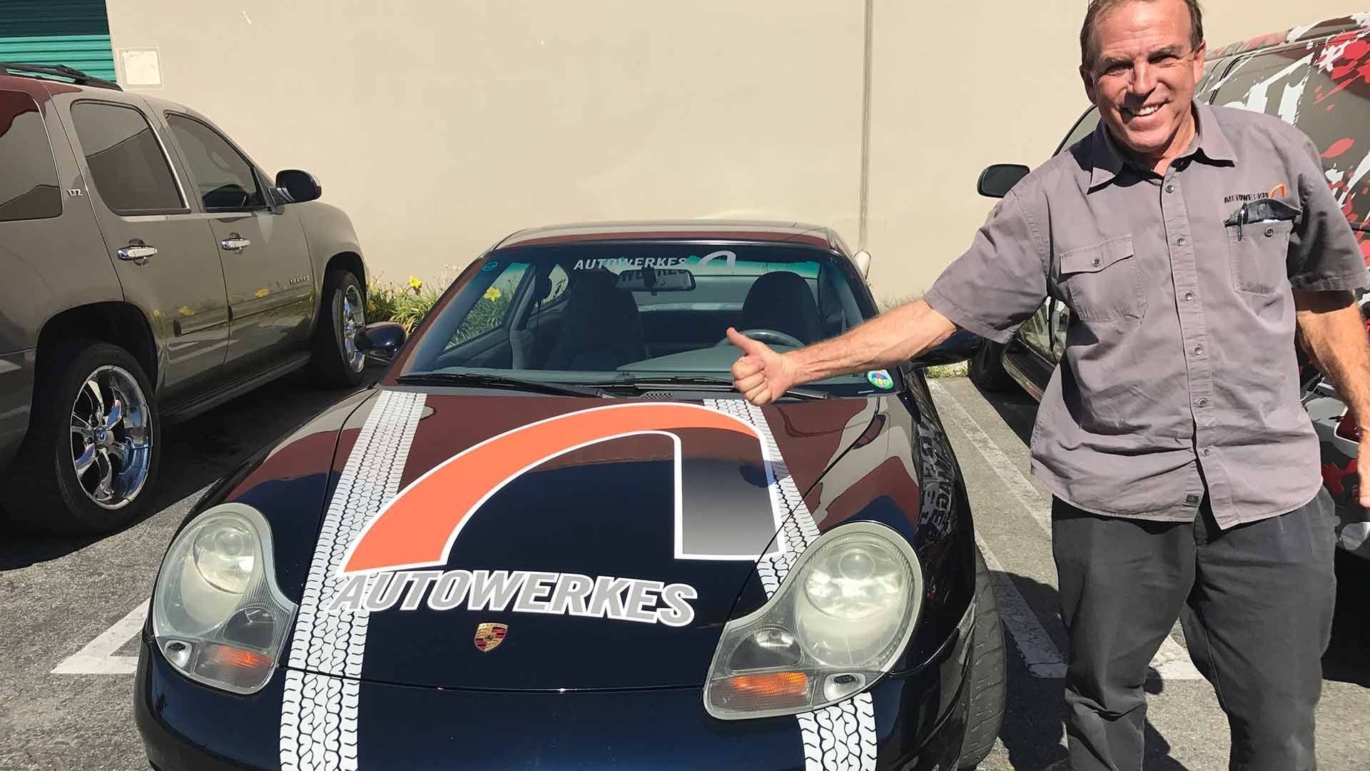 A man smiling and giving a thumbs up next to a black Porsche with a white and orange tire-track decal on the hood.