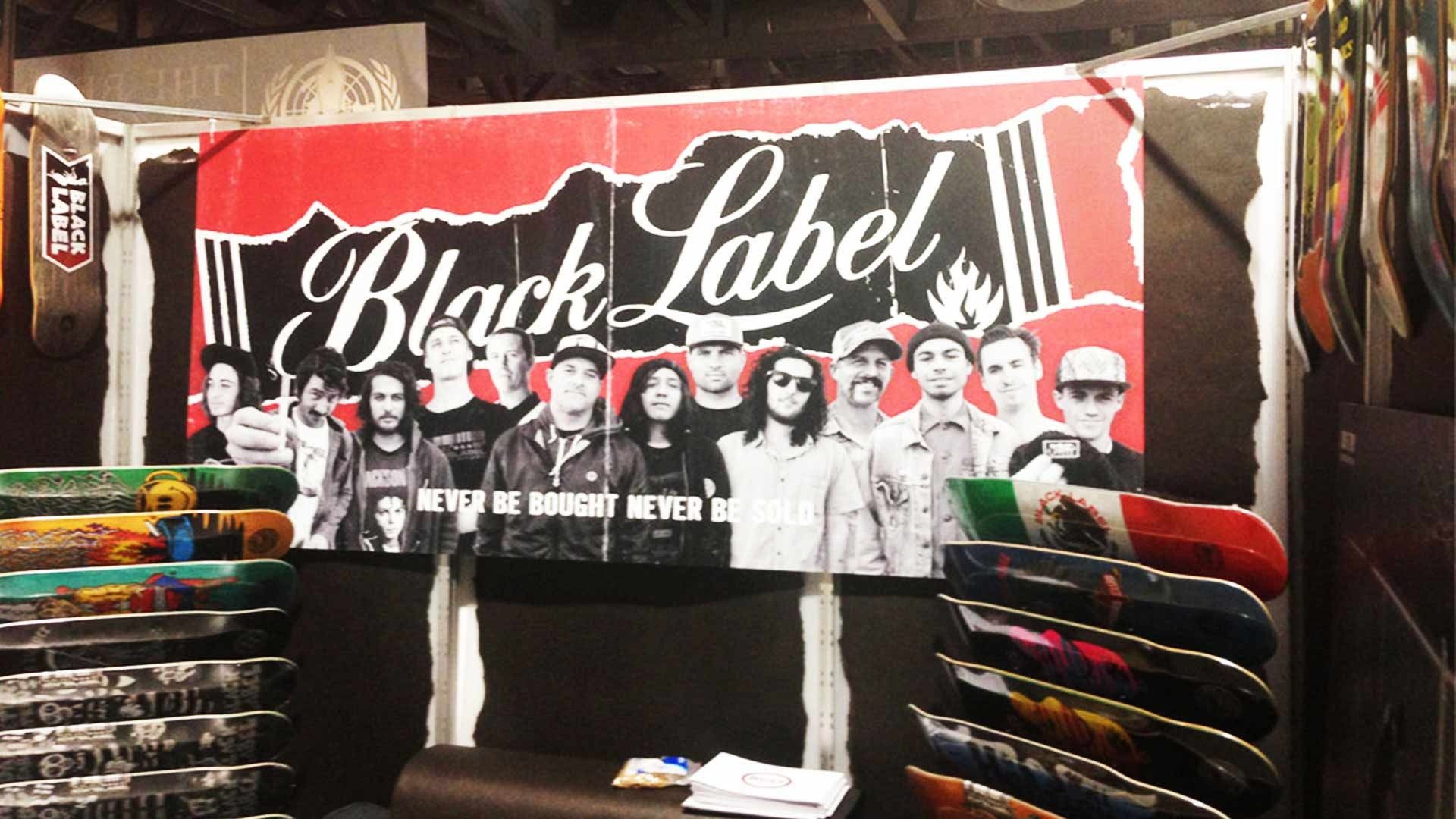 A Black Label skate brand trade show booth with a prominent logo, group photo, and racks of skateboard decks.