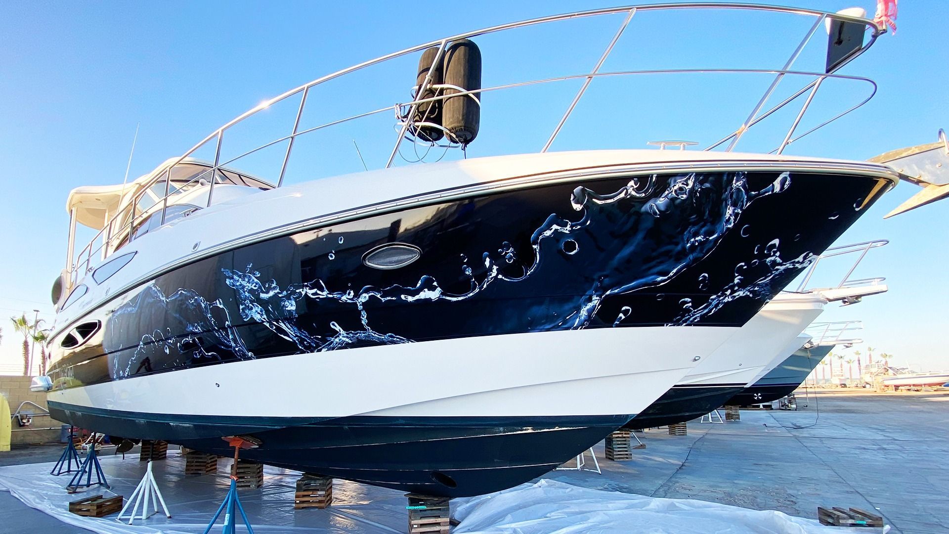 A white and black boat with a water-splash hull graphic, propped on stands in a dry dock area under a clear blue sky.